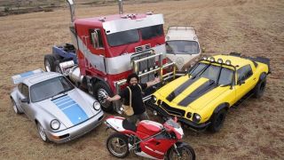 Still from the movie Transformers: Rise of the Beasts. Here we see a man standing in between 5 different vehicles. They are all Transformers in their vehicle disguise mode. There are 2 sports cars, a truck, a motorcycle, and a VW van.