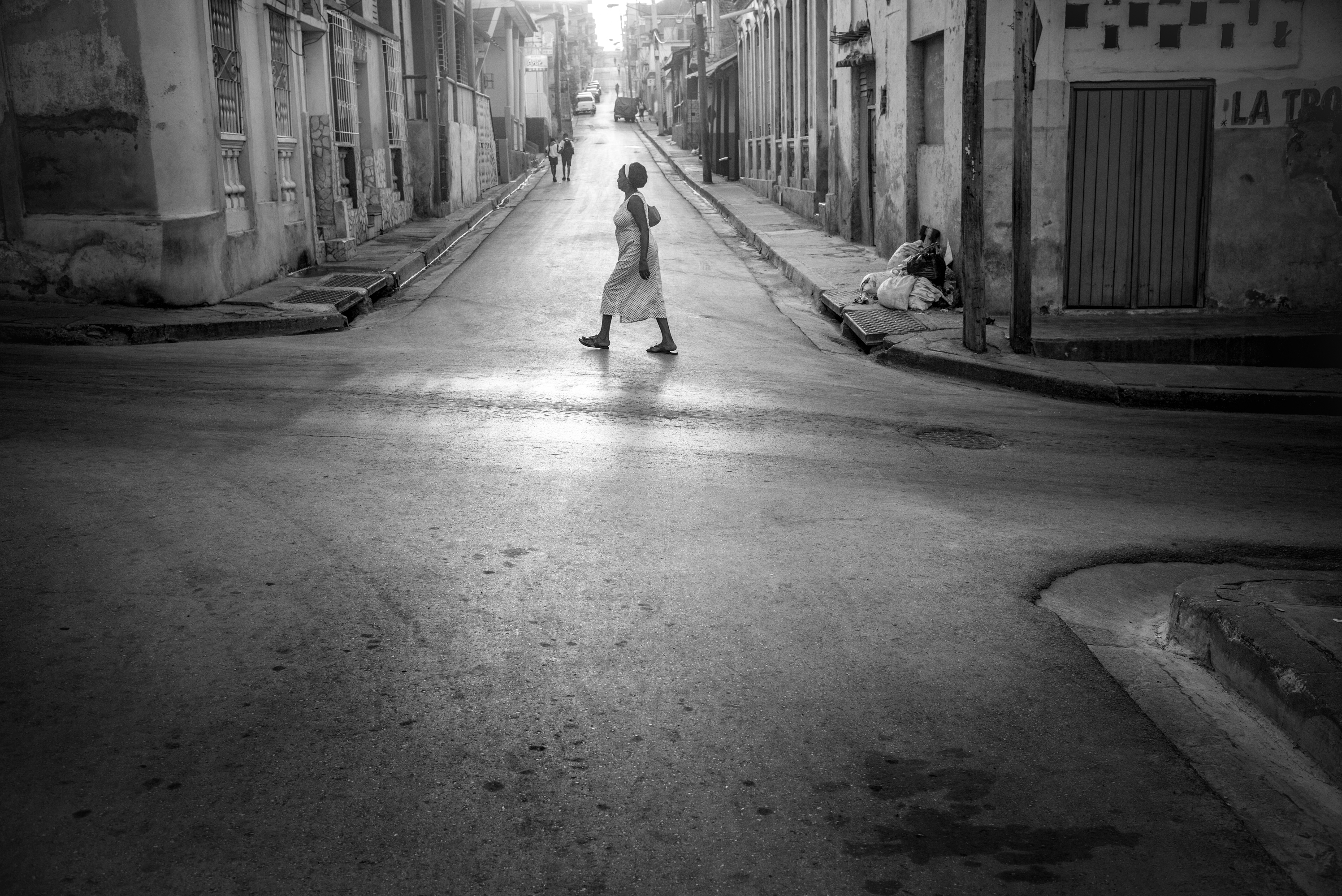 Black and white image of a woman in a light dress walking alone across a quiet, sunlit Havana street, her long shadow stretching across the wet tarmac.