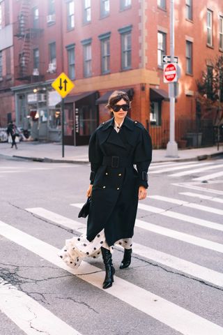 A woman walking in the street at New York Fashion Week wearing a puff sleeve dark trench coat with a black and white polka dot dress and black leather boots