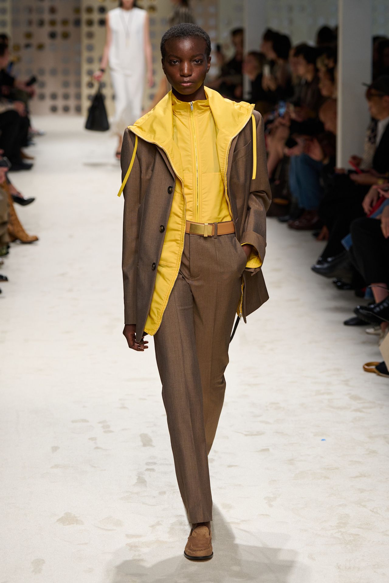 Layering staples for spring 2026 is shown in a photo from Tod's runway show, with a model walking while wearing a tan blazer over two yellow parka jackets, one tucked into tan trousers. The rest of the look is styled with a tan belt and tan loafers