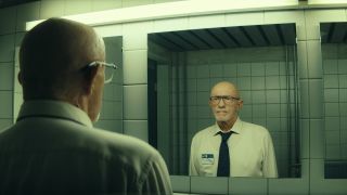 Bald man with glasses wearing a white shirt and black tie is starting intently into a public bathroom mirror.