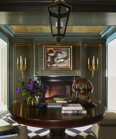 Cozy corner of entryway with dark green painted walls with gold trim, gold sconces on wall either side if a fireplace, large round wood table in foreground decorated with books and ornaments
