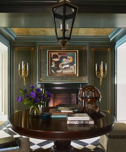 Cozy corner of entryway with dark green painted walls with gold trim, gold sconces on wall either side if a fireplace, large round wood table in foreground decorated with books and ornaments