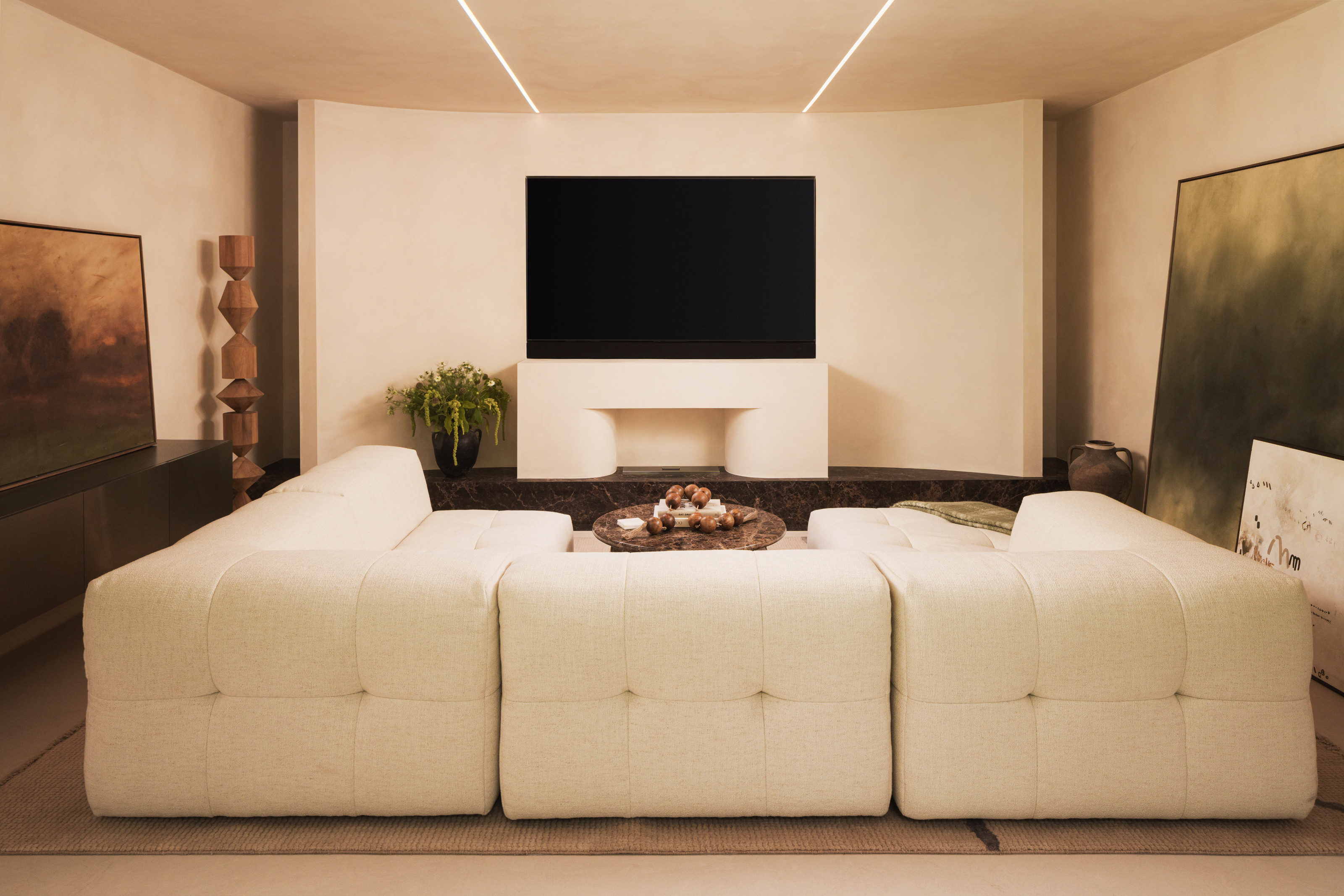 Media room with beige textured walls, cream U-shaped modular sofa, cream console built into a brown marble base with a TV on top, and brown marble coffee table
