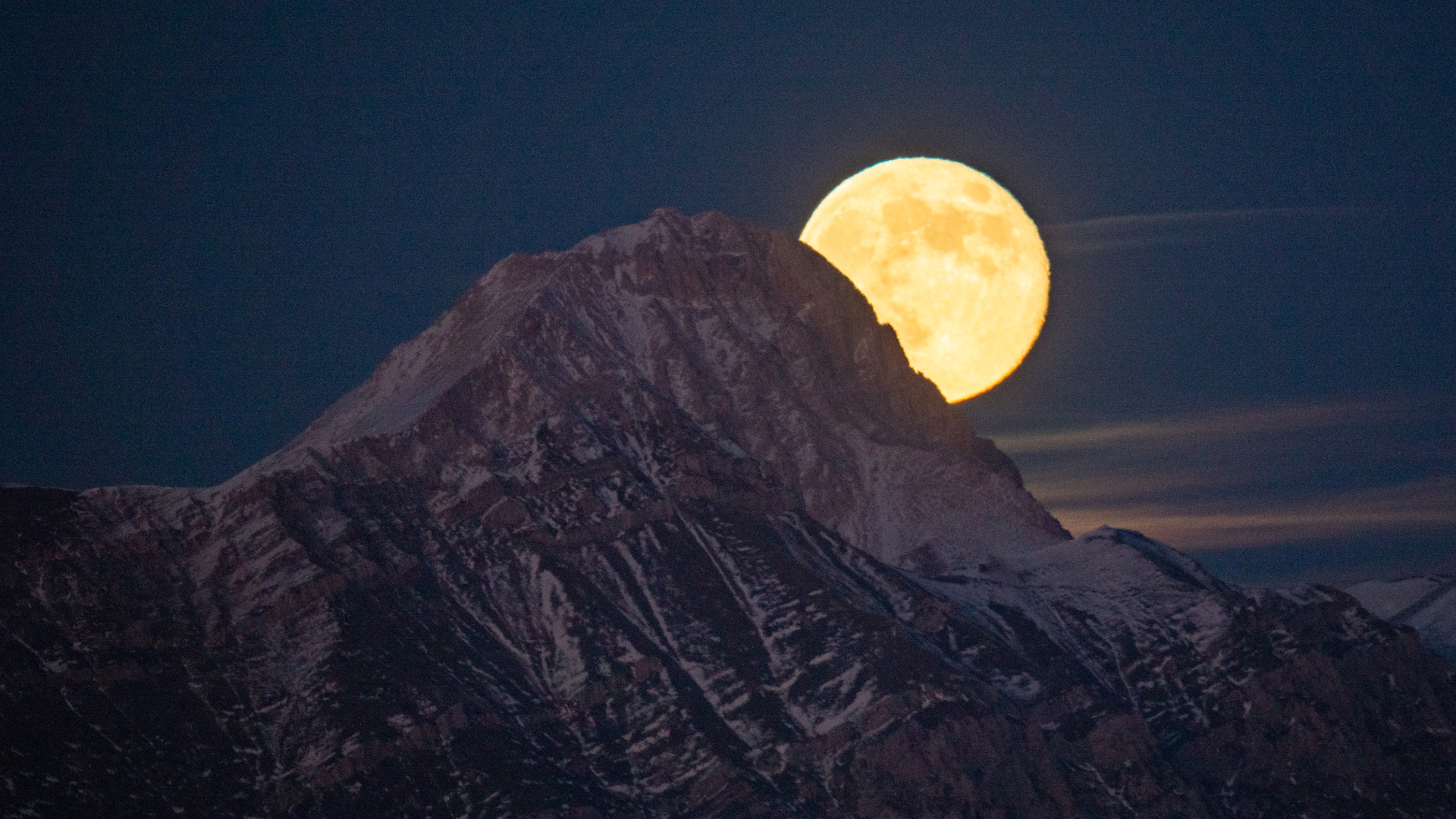 A yellow full moon is pictured rising to the right of a snow-dappled mountain peak against a dark blue sky.