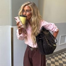 Influencer @emilisindlev sips on a coffee wearing a pink shirt tucked into dark brown trousers.