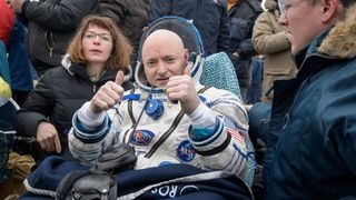 a bald white astronaut in a pressure suit gives two thumbs up as he sits in a chair outside surrounded by people in winter jackets