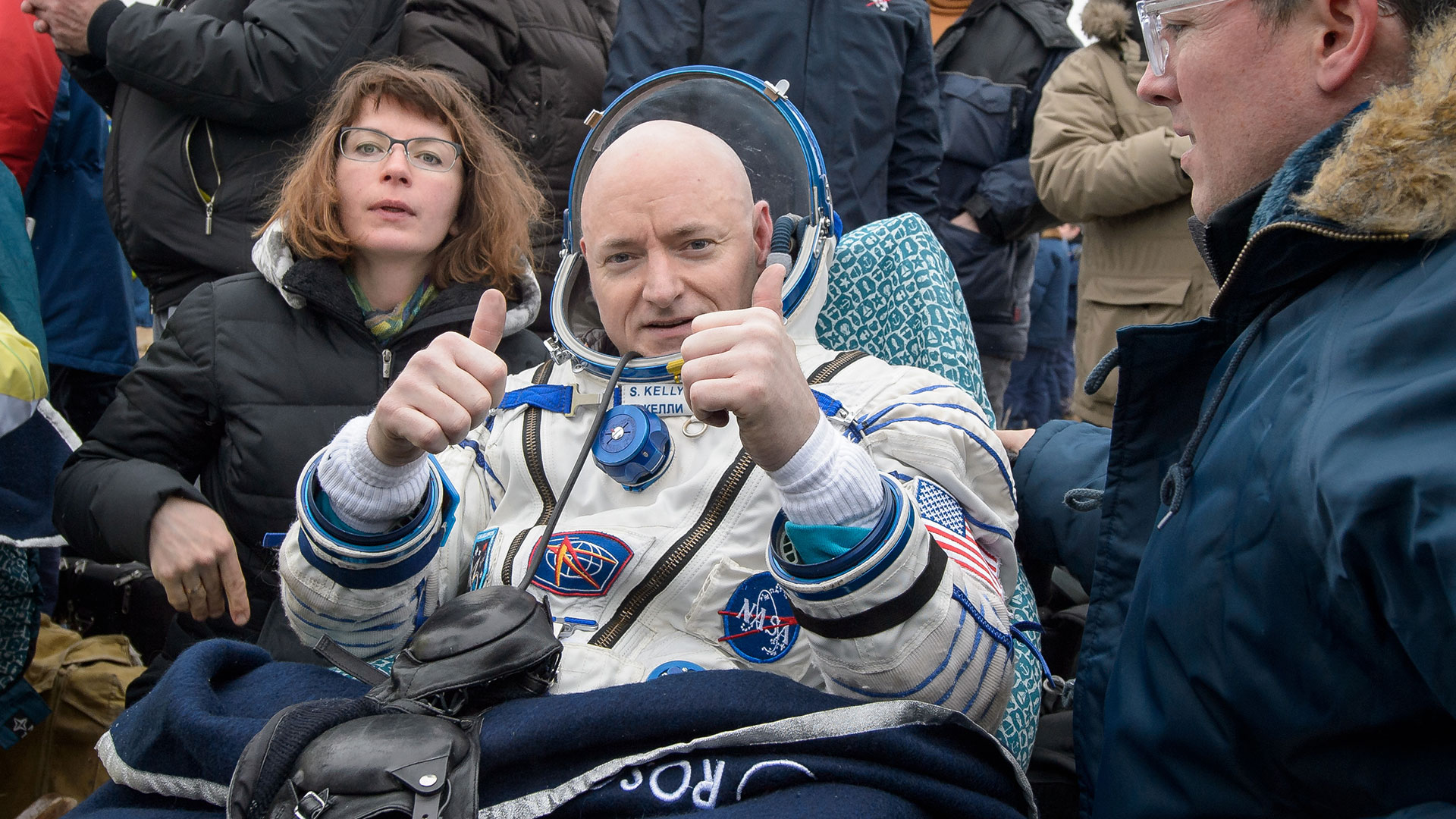 a bald white astronaut in a pressure suit gives two thumbs up as he sits in a chair outside surrounded by people in winter jackets