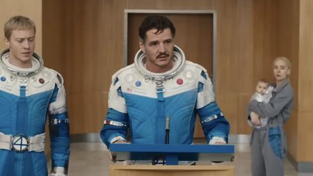 Reed Richards speaking into microphone during press conference, with Sue in the background holding Franklin in The Fantastic Four: First Steps