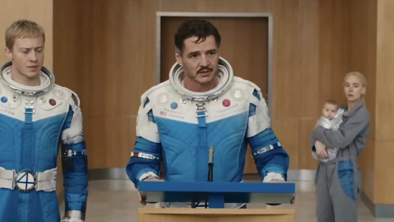 Reed Richards speaking into microphone during press conference, with Sue in the background holding Franklin in The Fantastic Four: First Steps