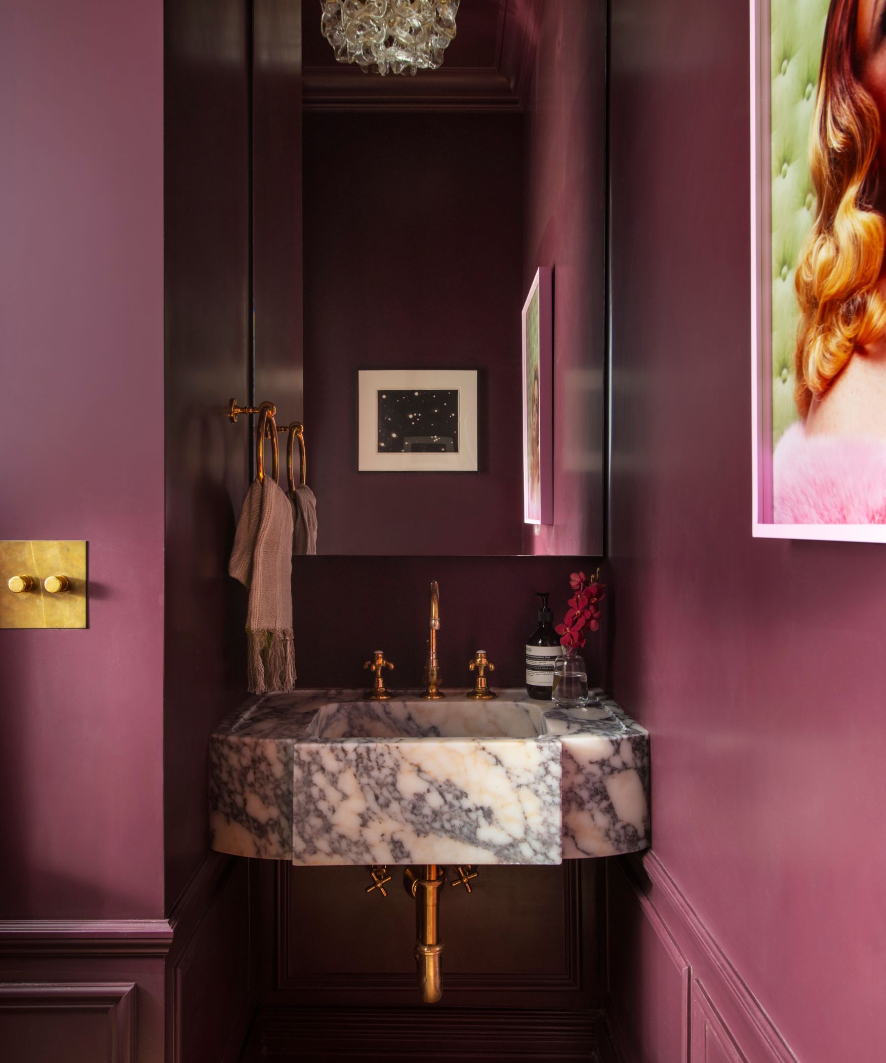 Small bathroom with marble sink, aubergine painted walls, gold faucet and towel ring and a framed picture on right side of wall
