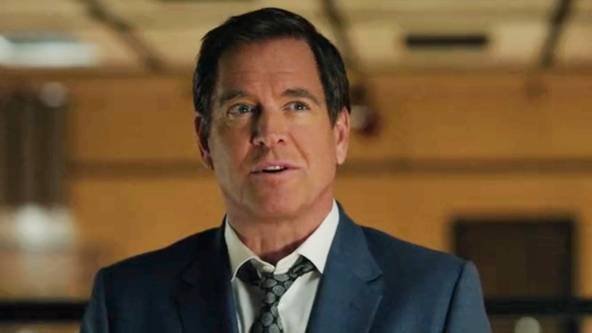 Michael Weatherly with the orange walls in the background in the NCIS Season 24 premiere. He has on a blue suit and tie.
