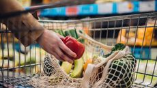 A woman puts a red bell pepper in a mesh bag in a grocery cart.