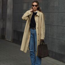 woman wearing khaki trench coat, blue jeans, and carrying brown suede purse
