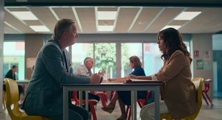 Daniel (Douglas Henshall) and Erin (Evin Ahmad) sit on opposite sides of a table at the school, having a meeting. Other pairings having similar meetings are visible behind them.