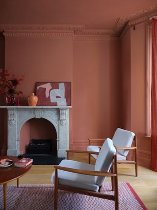 A living room with Farrow & Ball’s Red Earth matt paint on walls and ceiling, with a white marble fireplace and white/teak armchairs