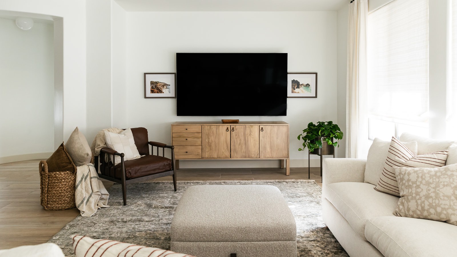 Living room with TV mounted on wall above retro wooden media unit, with cream sofa and footstool pictured in foreground. Also pictured are a brown armchair, miscellaneous cushions, a potted plant, and two framed pictures, in a room with white walls and curtains.