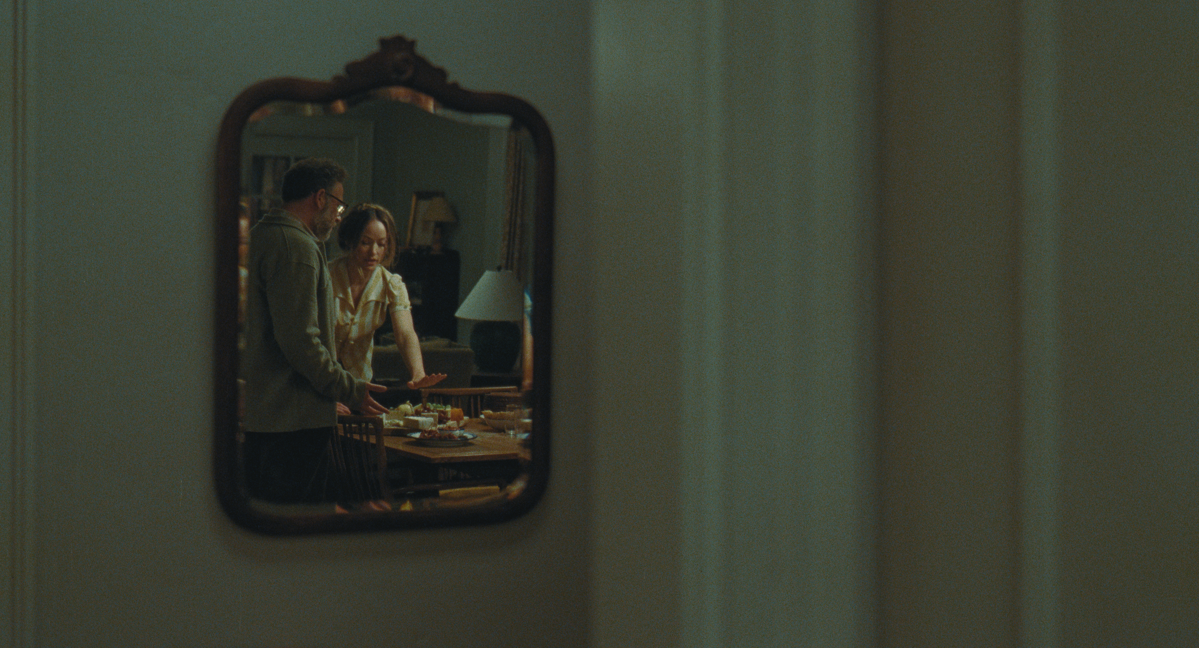 a shot of seth as joe and olivia wilde as angela in a hallway mirror as they prep food in the kitchen in a still from the invite