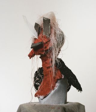 Sculpture, red clay head, black feathers, dried seaweed, and broken Japanese pottery pieces, sat on a soft grey covered plinth, white background