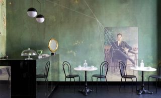 A restaurant with round tables, black chairs, a black serving counter, round pendant lights, green walls and a wall painting of a seated man in a suite.
