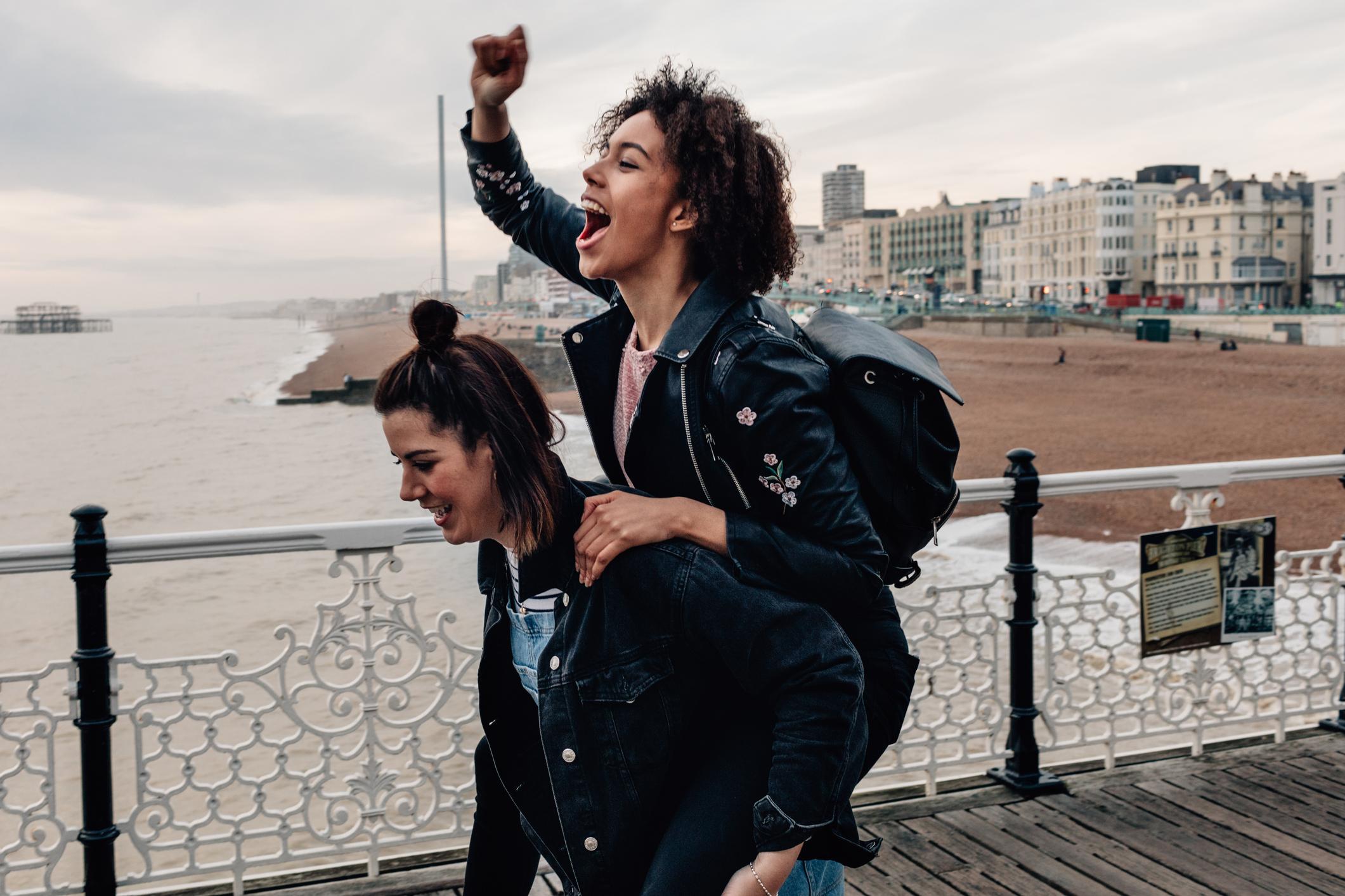  One woman is carrying another woman on her back while they have fun at the seaside.  