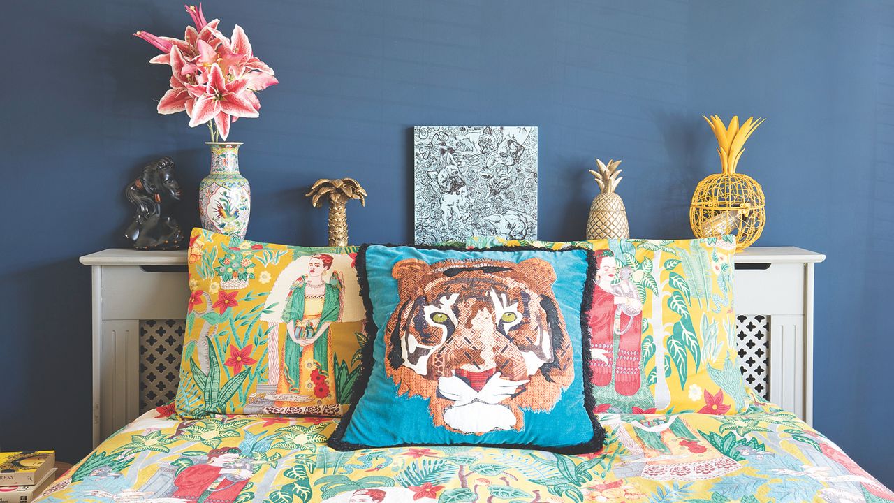 blue bedroom with bright bedding, lion cushion, artwork, flowers, radiator cover behind and used as a shelf 