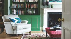 A green living room seen through a white open door, with green shelving, green and white wallpaper and armchair, light wood flooring, brightly patterned rug, dark pink ottoman, and white mantelpiece.