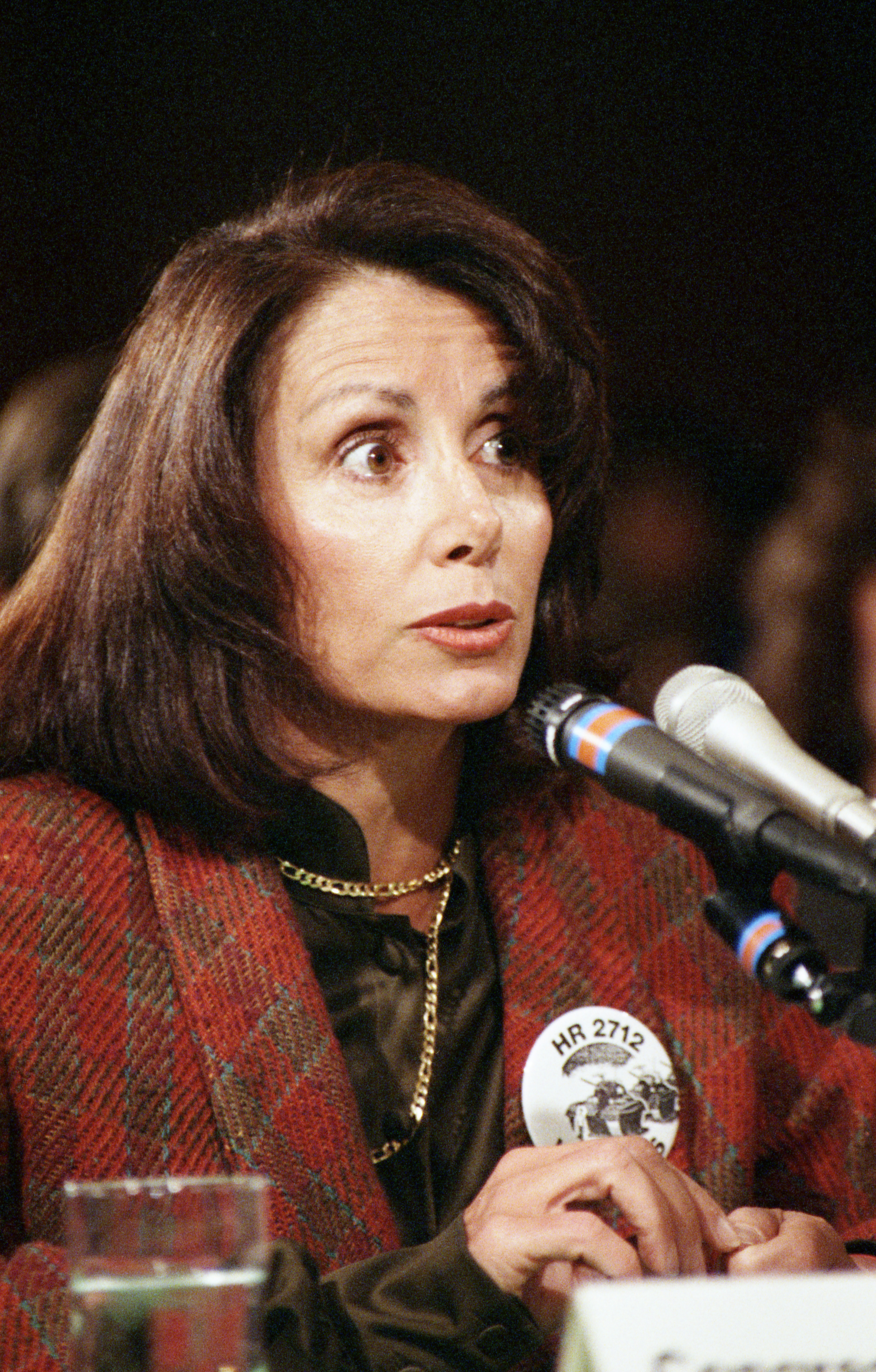 An older photo of Nancy Pelosi speaking into two microphones, wearing a red patterned jacket over a black blouse.