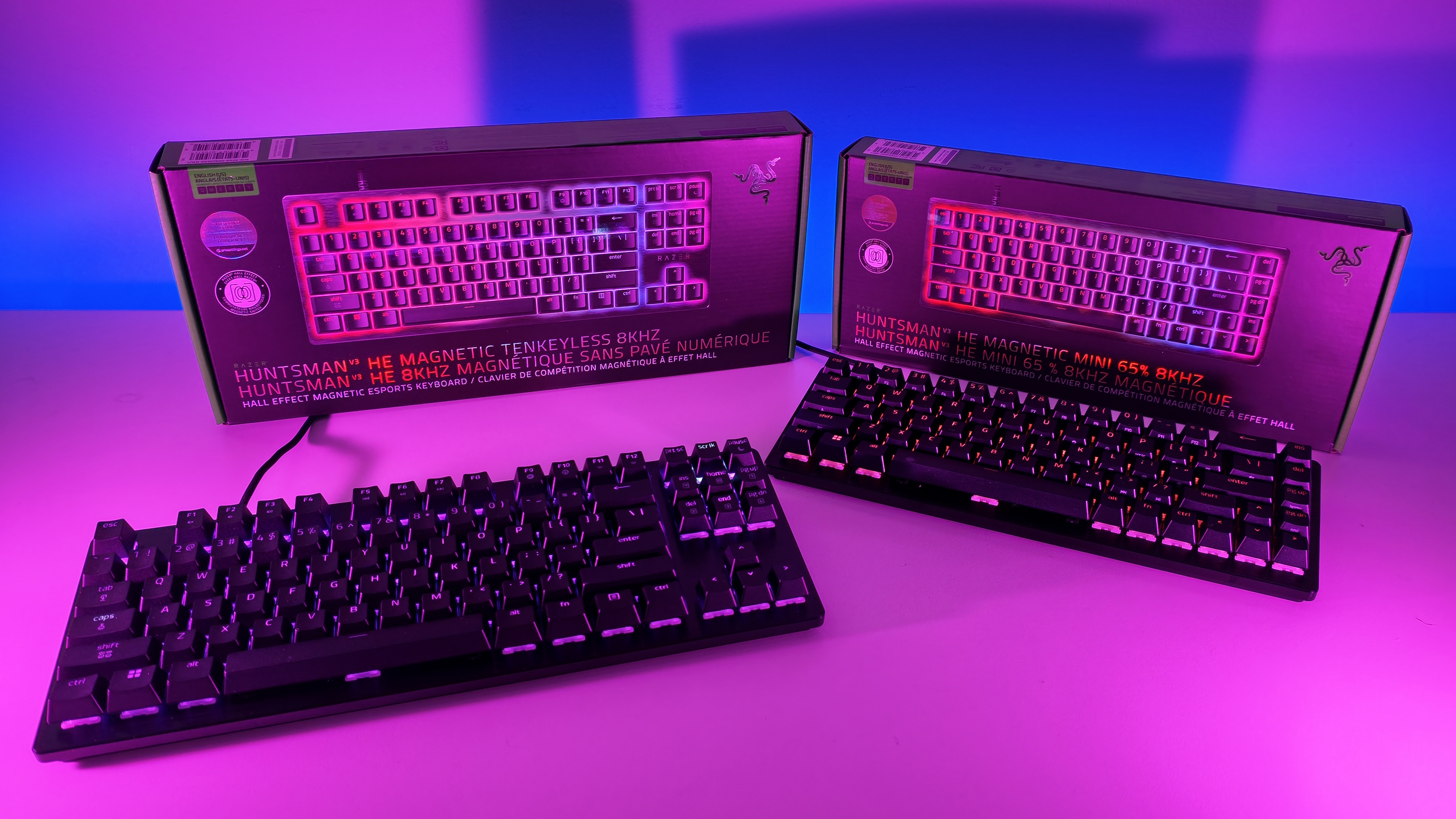 Low-angle shot of Razer Huntsman V3 HE TKL and Mini 65% keyboards with RGB backlighting placed in front of their retail packaging.