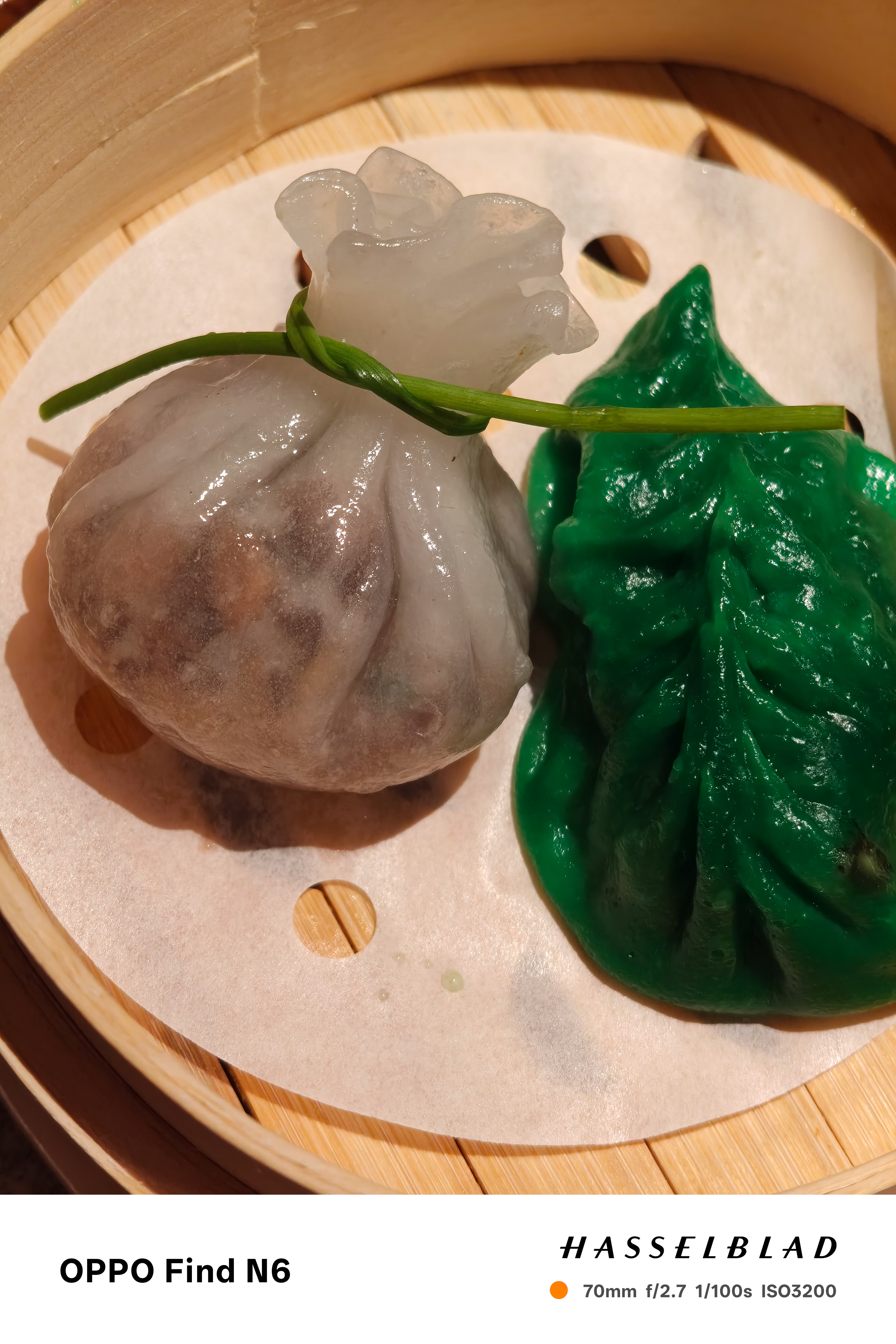 A close-up of two dumplings in a bamboo steamer. One is a translucent white "money bag" style dumpling tied with a green chive, and the other is a vibrant green, leaf-shaped dumpling with intricate pleating.