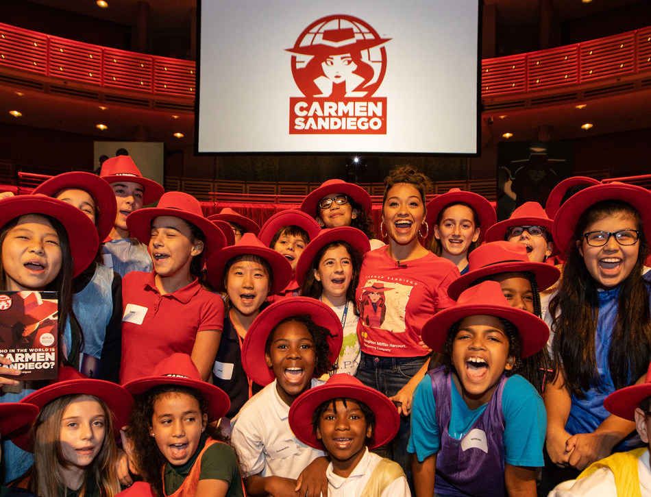 Kids wearing Carmen Sandiego gear and red hats smile and laugh under a banner displaying her logo