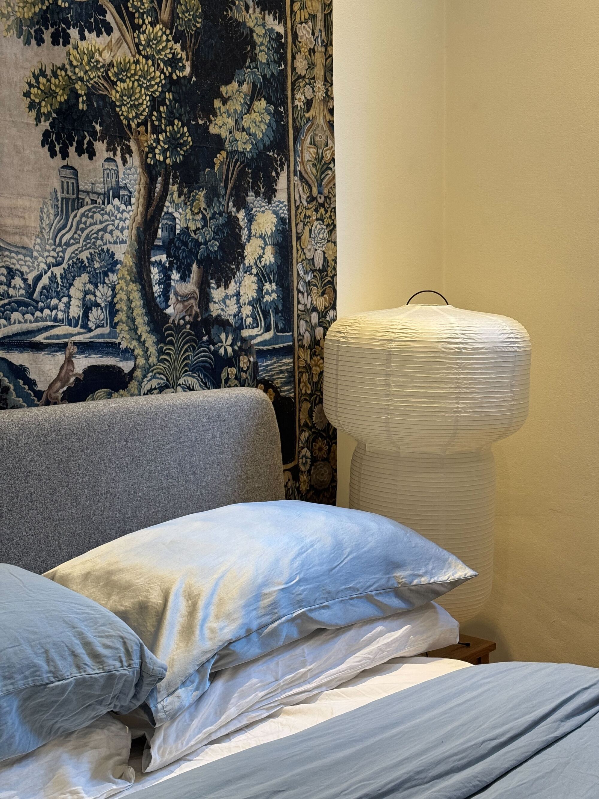 A close up of a bed with blue and white sheets, a paper lantern next to the bed, a tapestry hanging above a gray headboard, and a blue silk pillowcase
