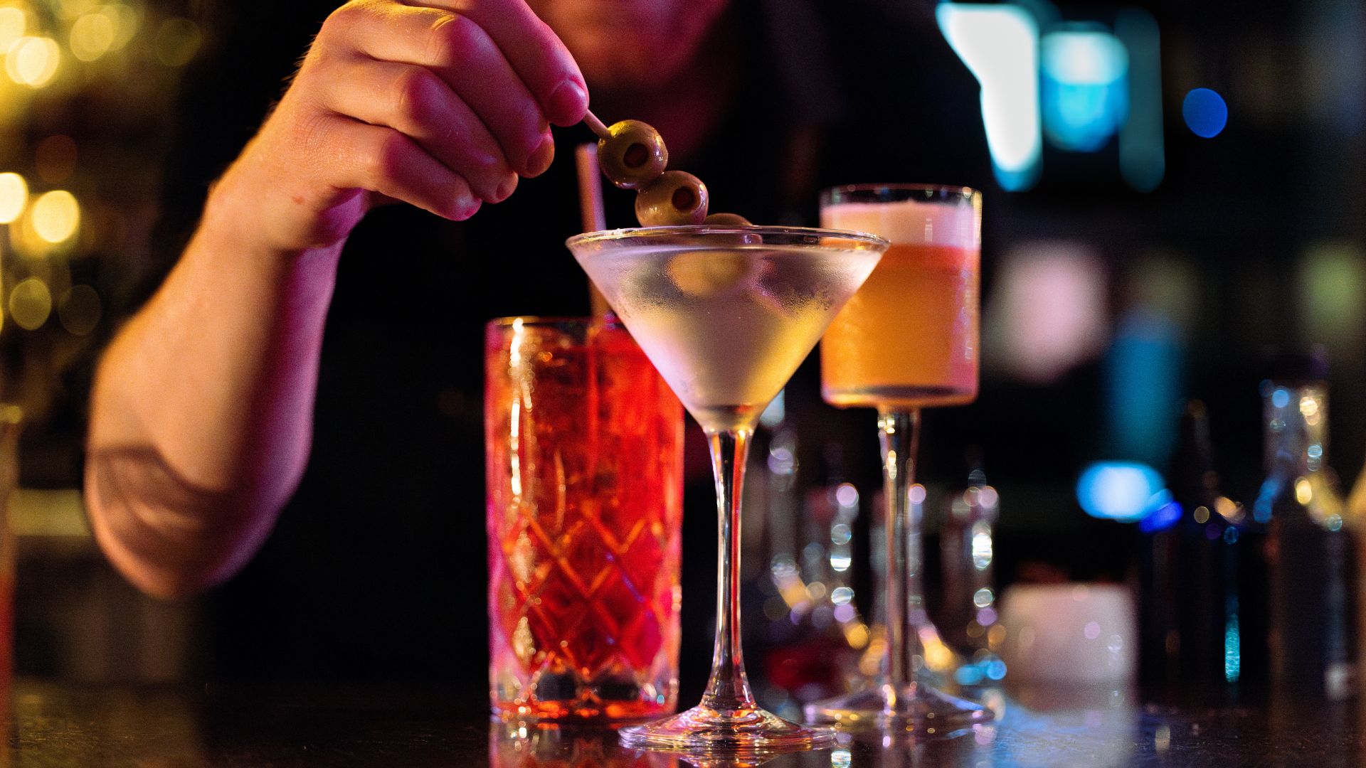 In the foreground a hand places a toothpick with three olives into a martini glass in a darkly lit bar. Behind are two other vibrant cocktails, and out of focus in the background is the rest of the bar lit in purples, yellows, and blues. The three glasses gleam on the counter.