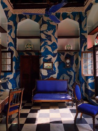 Traditional sitting room drenched in patterns and Indigo. The floors is black-and-white checkered tile, and the walls are an artistic blue and white wallpaper of arm-looking figures. The furniture is traditional style wood pieces with indigo upholstery fabric.