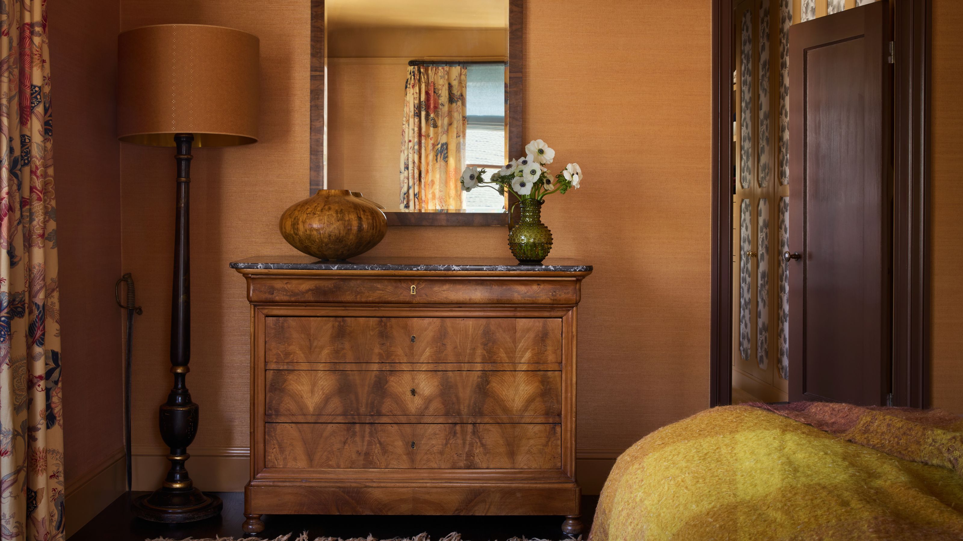 a caramel colored wallpaper primary bedroom with art deco antique wooden furniture and floral drapes
