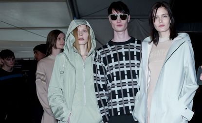 A row of three models pose for a picture with some people behind