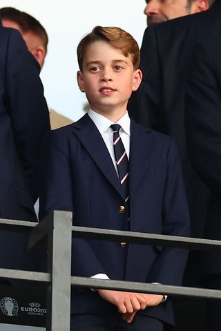 Prince George wearing a blue suit and striped tie standing up at a stadium