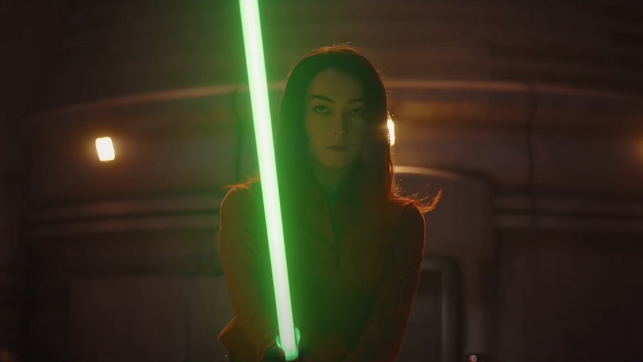 Still from the Star Wars T.V. Show Star Wars The Mandalorian. Here we see Sabine Wren with brown eyes and long dark, shoulder-length hair. She is wielding a green lightsaber, ready to fight.
