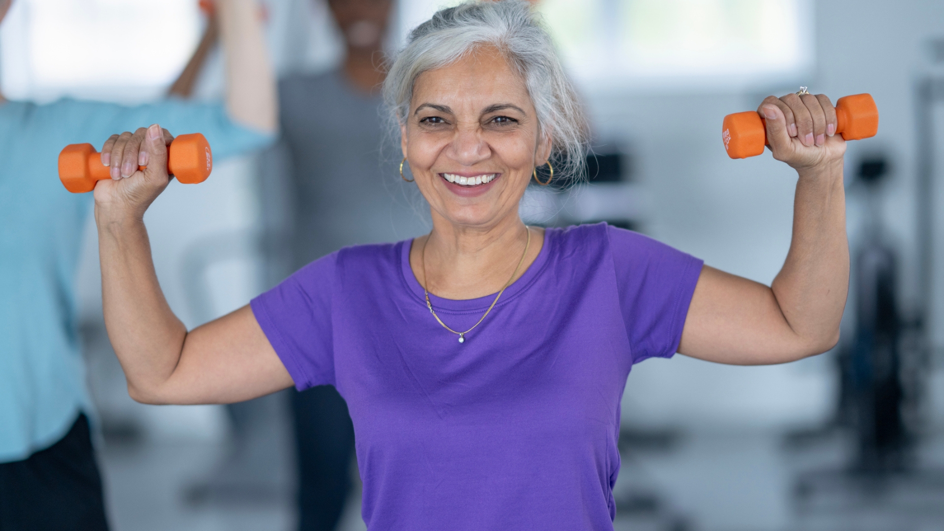 woman wearing a purple tshirt facing the camera, smiling and holding a small orange dumbbell in each hand at shoulder height half way through an overhead press. 