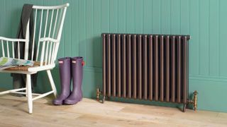 a Bisque Tetro aluminium radiator in a hallway, with a green wall, wooden floor, and a chair and wellies next to it