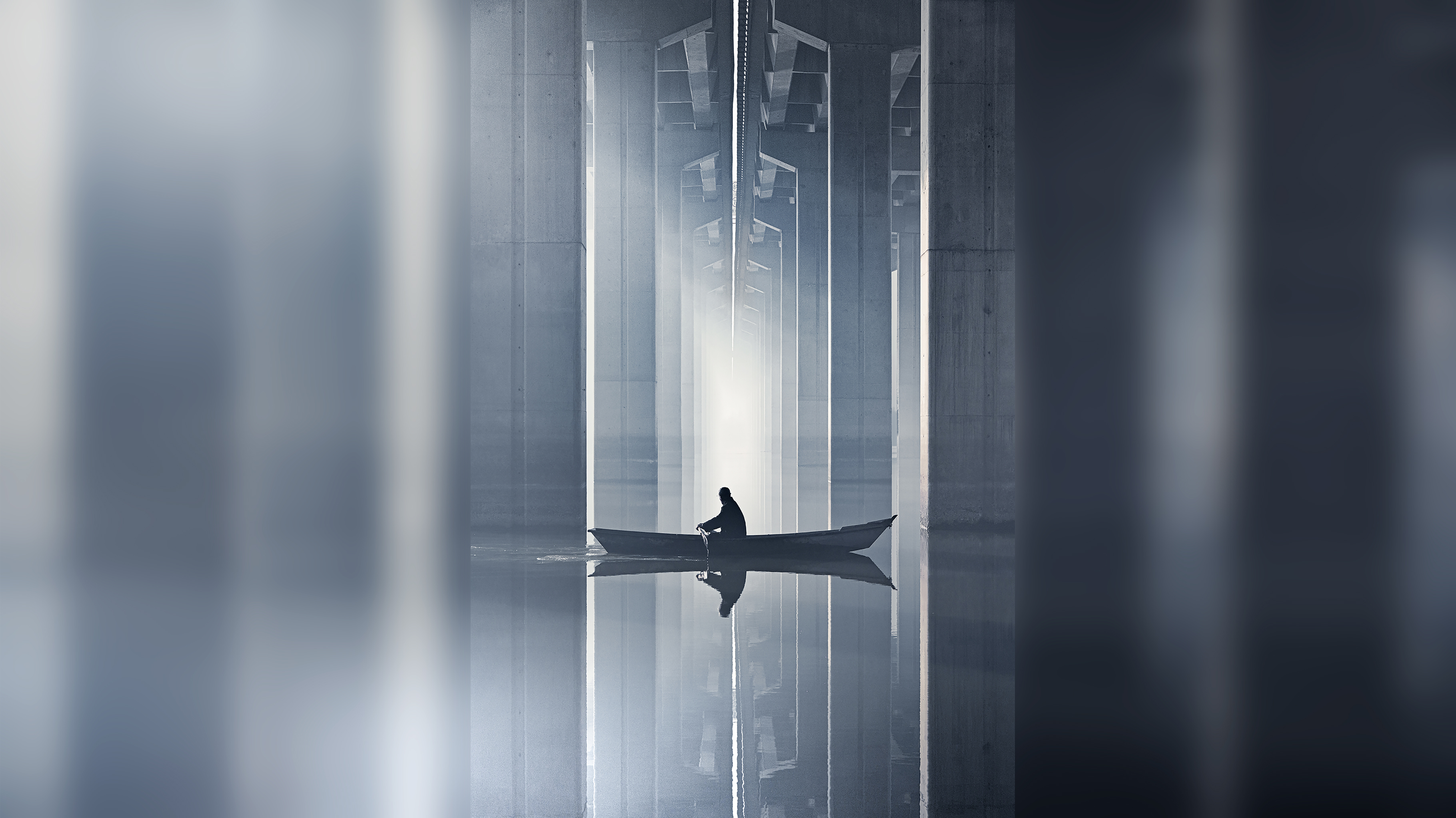 A person rows a small boat through calm waters under a modern bridge, with towering concrete beams