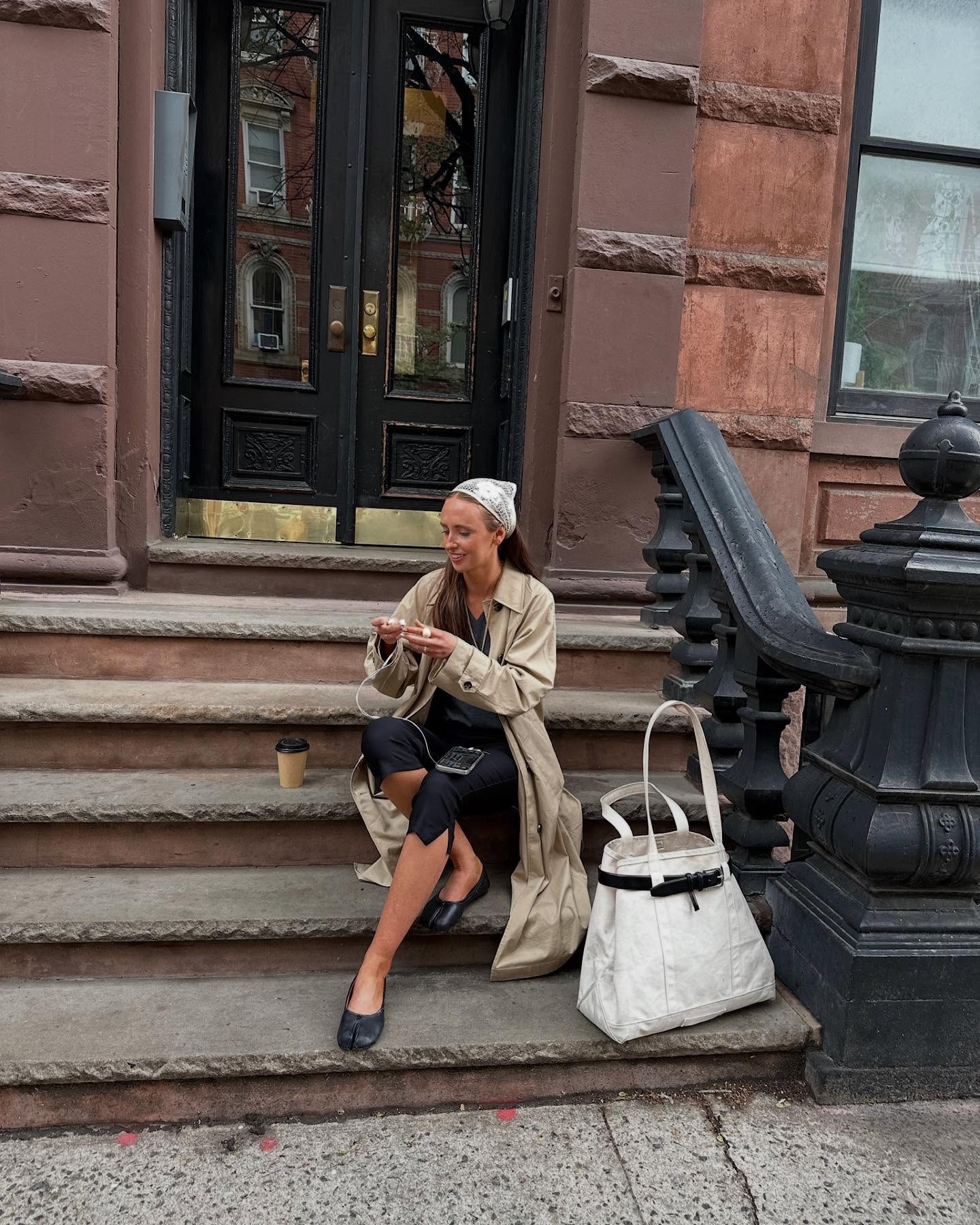A woman sitting on a stoop wearing black tabi flats, black capris, a trench coat, and a lace headscarf.