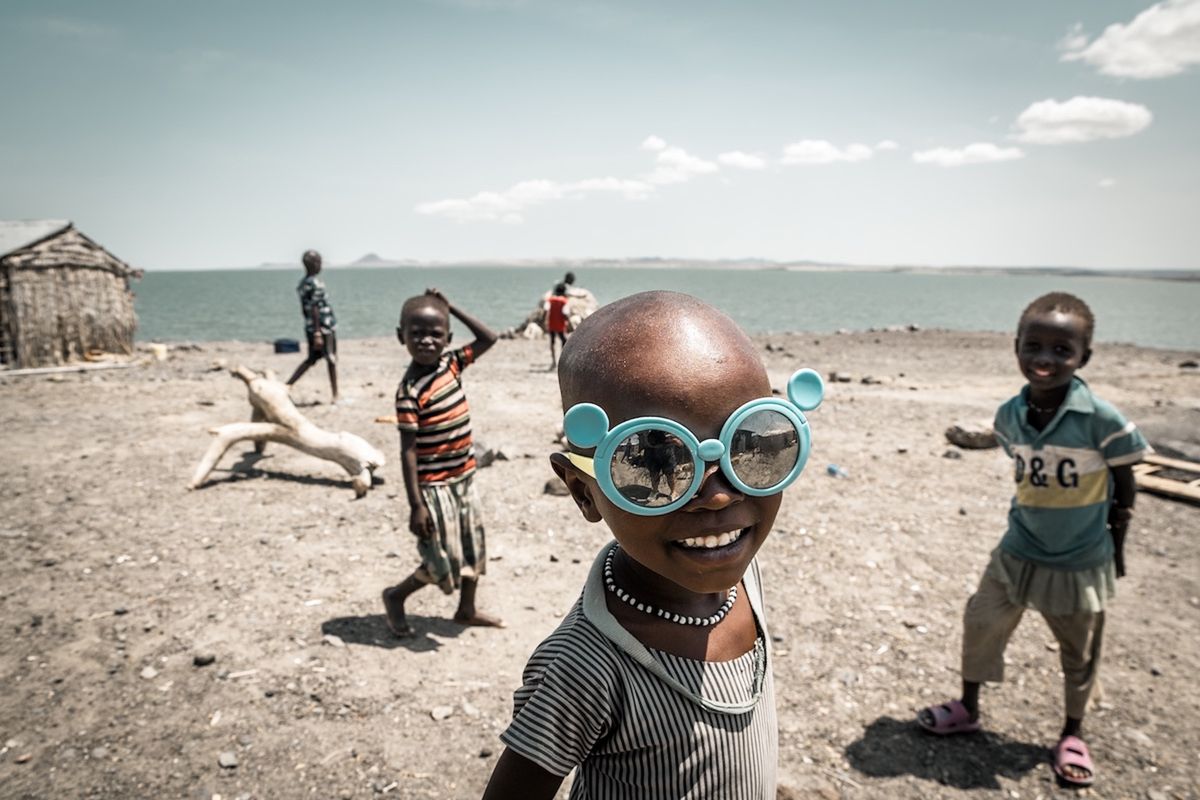 children playing by the coast with the sea in the background and the children in the foreground is wearing flamboyant blue sunglasses