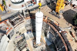aerial view of a crane lowering a large white cylindrical rocket motor into place in a big, unfinished building.