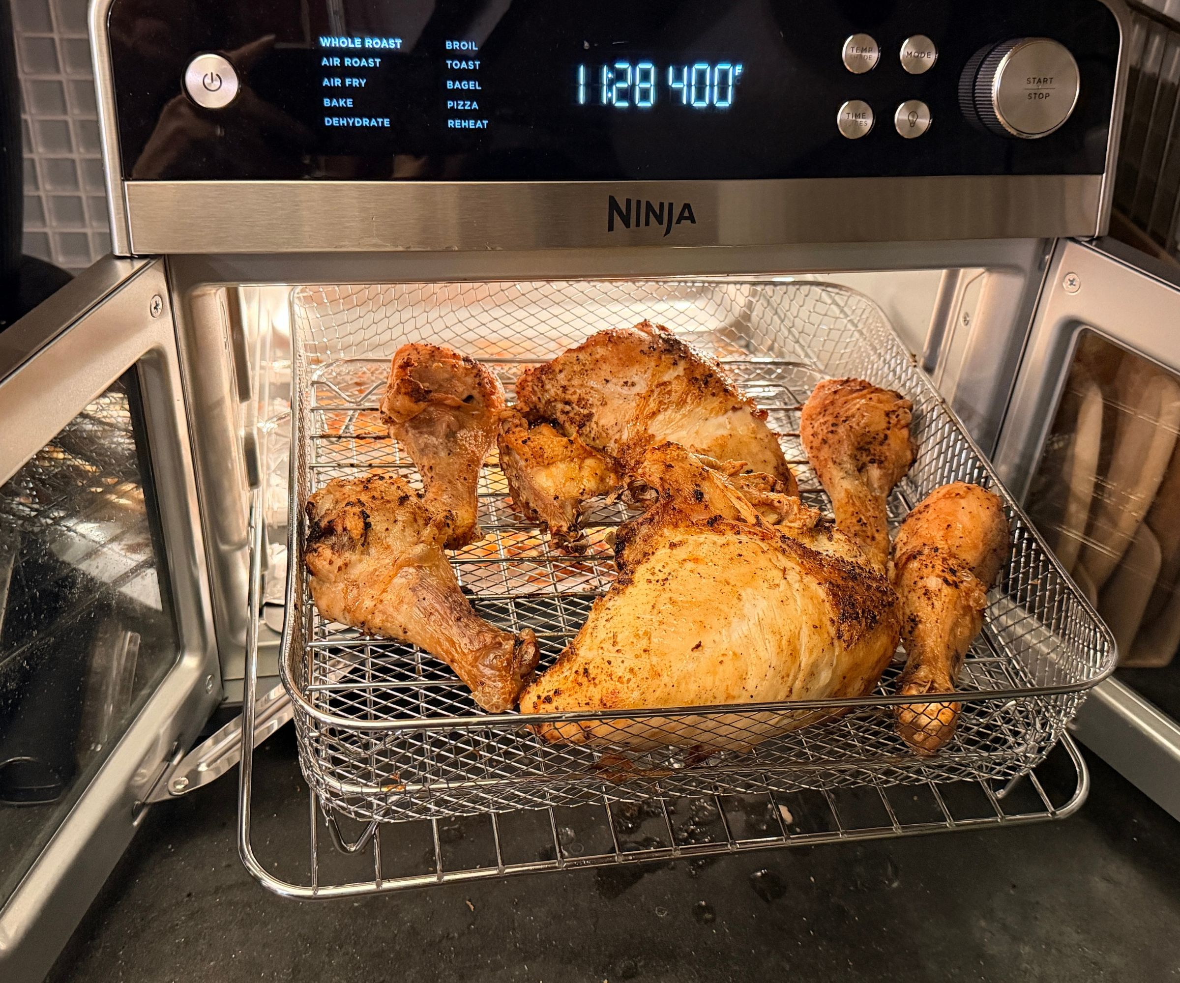 Cooked chicken breasts and legs in a wire basket in the open double-door Ninja French Door Premier air fryer on a black kitchen counter