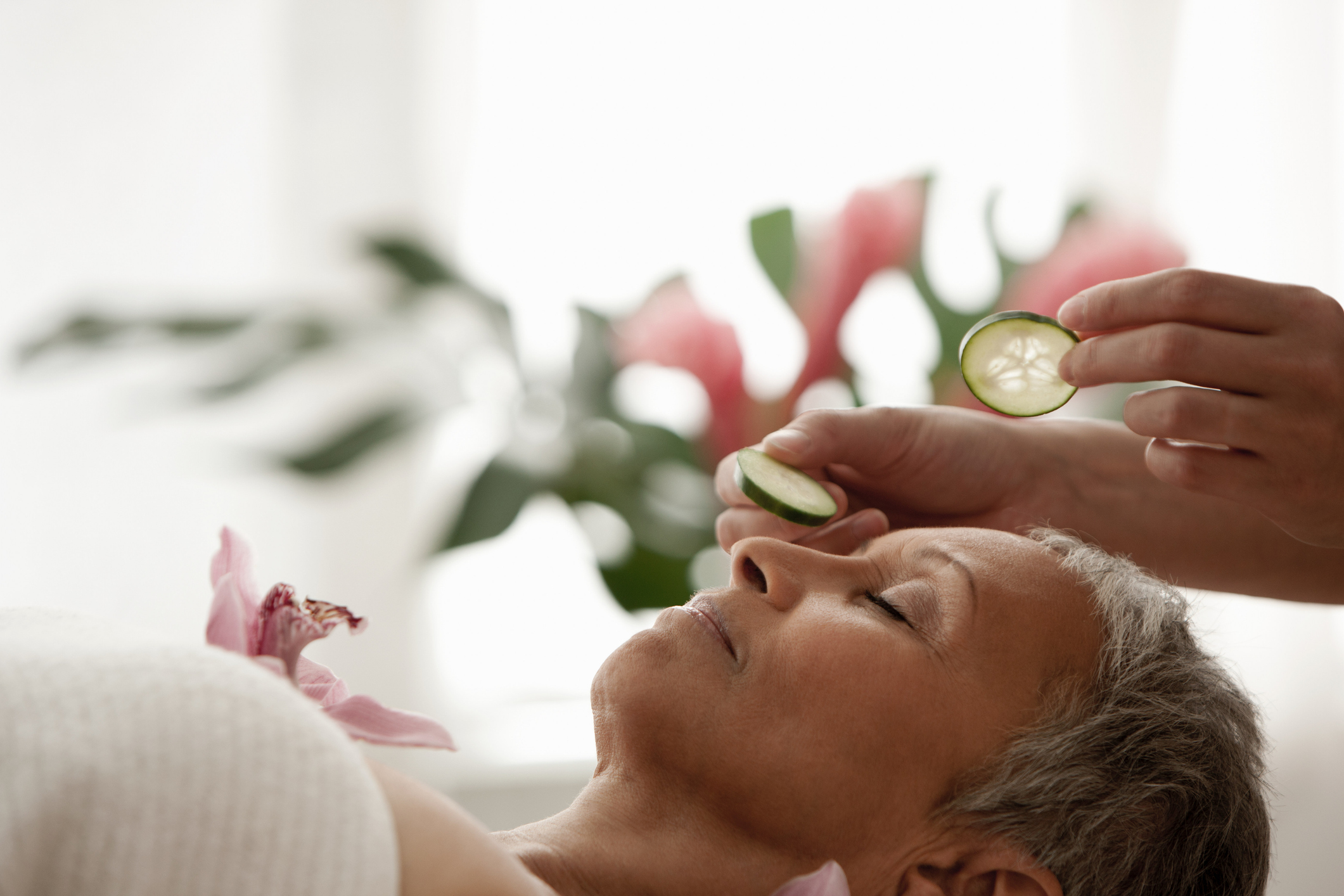 An older woman rests on a massage table at a spa. The massage therapist is about to put cucumber slices on her eyes.