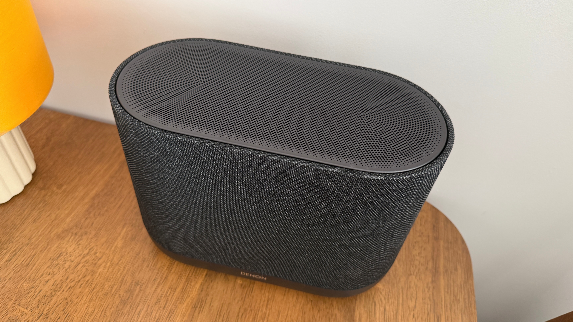 A top-down view of the Denon Home 400 wireless speaker on a wooden table. A lamp is just visible to the left.