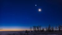 The moon shines in the evening sky with a bright star-like planet glowing close to its upper left above a snowy field lined with trees. The glow of the setting sun can be seen brightening the horizon.