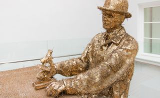 A sculpture covered in a golden sequence. A man, wearing a hat, looks at a microscope on the table.
