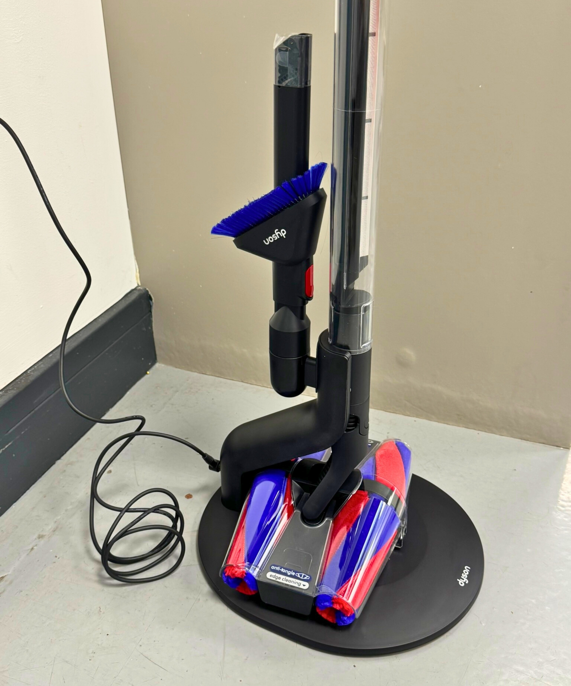 The Dyson PencilVac Fluffycones vacuum on its charging base with attachments attached at the back, with cable running to the left, on gray floor in front of gray wall.